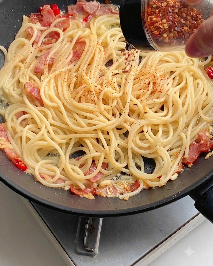 Rotes Chilipulver wird gleichmäßig über Spaghetti in einer Pfanne gestreut.