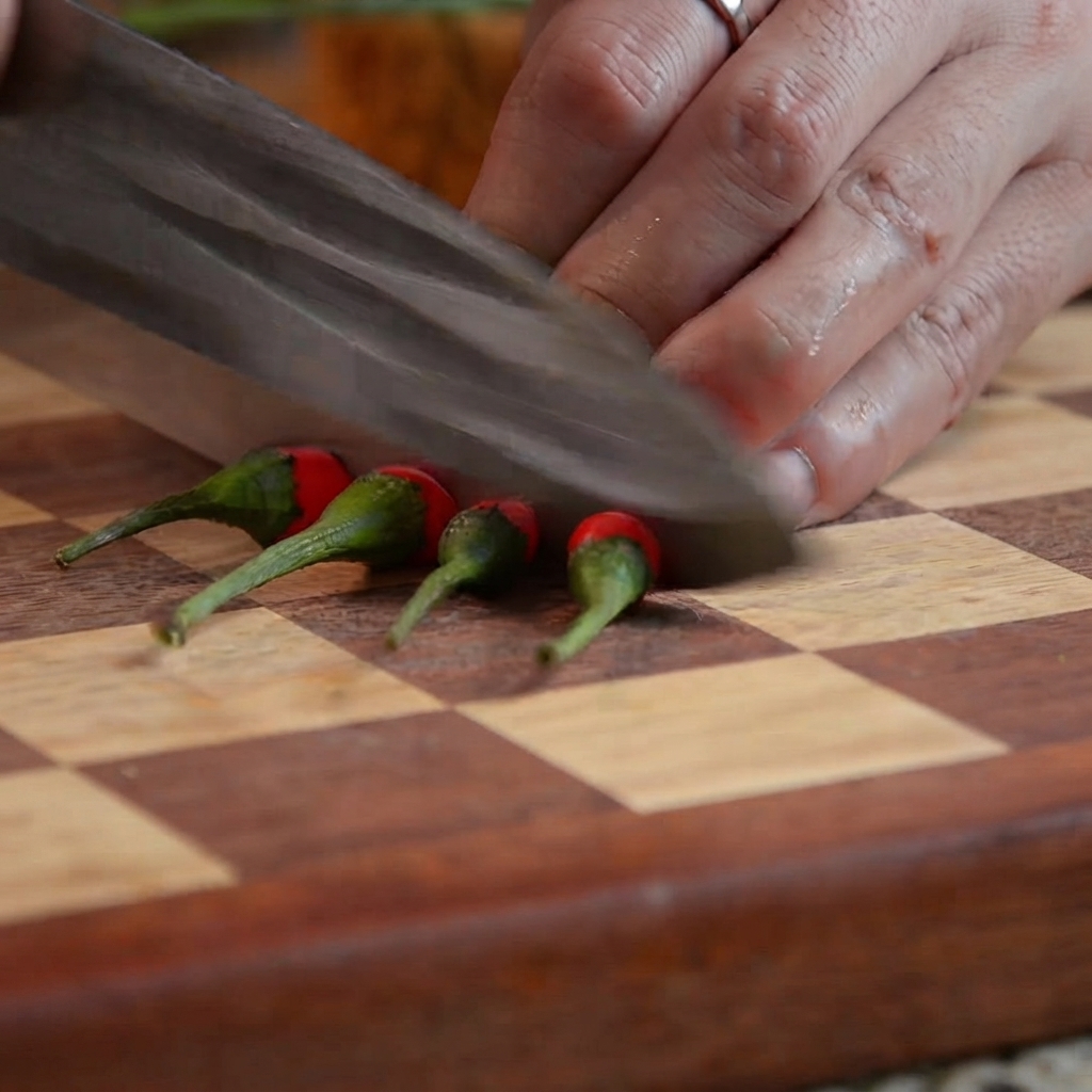 Ein Küchenmesser schneidet rote Chilischoten auf einem Holzschneidebrett in kleine Stücke.