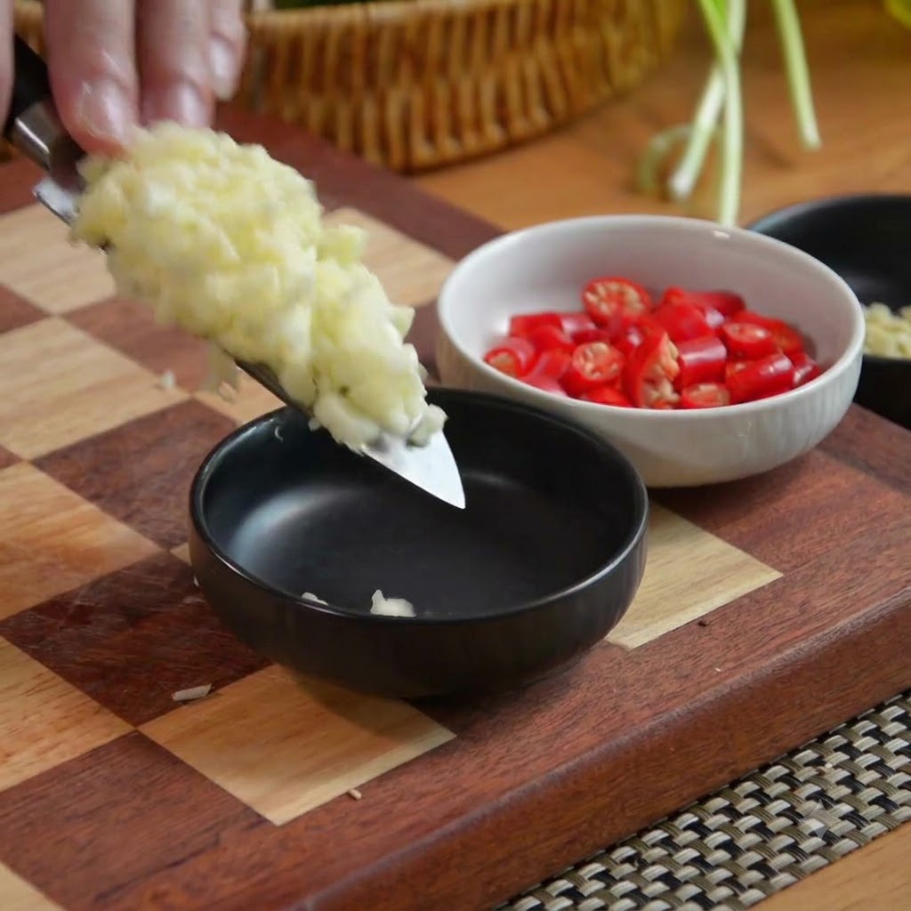 Gehackter Knoblauch auf einer Messerklinge wird zusammen mit einer Schüssel gehackter roter Chilis in eine schwarze Schüssel gegeben.