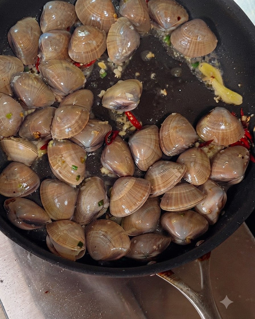 Weißwein wird aus einer Flasche in eine brutzelnde Pfanne mit Muscheln, Knoblauch und Chilischoten gegossen.