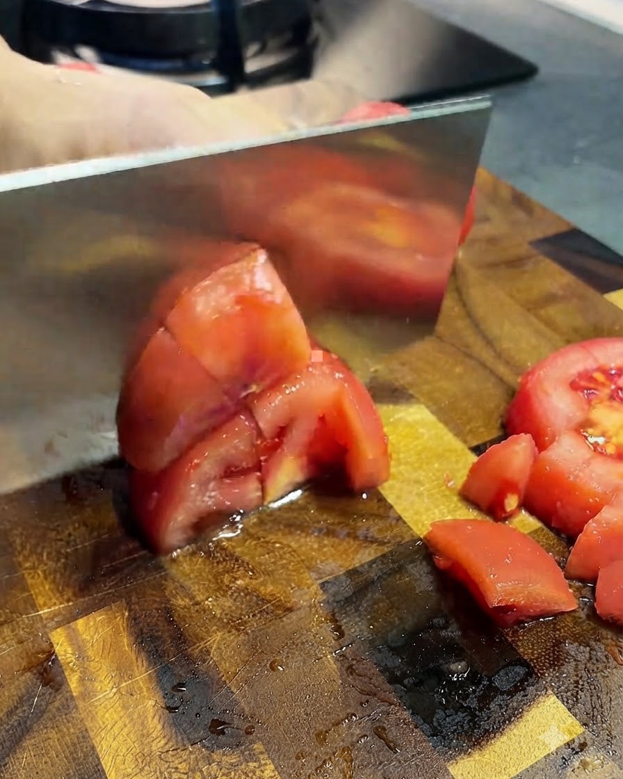 Geschälte Tomaten werden mit einem Hackmesser auf einem Holzbrett in kleine Stücke geschnitten.