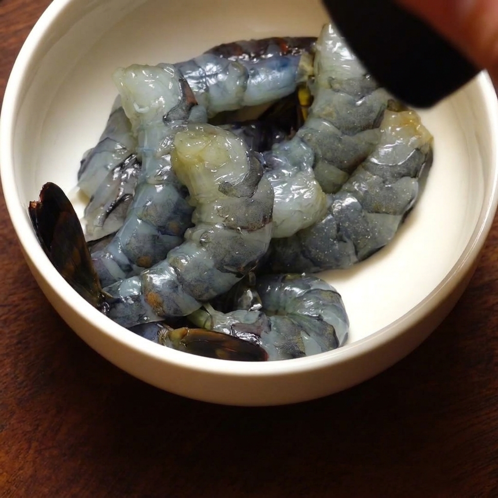 Eine Schüssel mit geschälten Black-Tiger-Garnelen, die mit frisch gemahlenem schwarzen Pfeffer aus einer Mühle gewürzt werden.