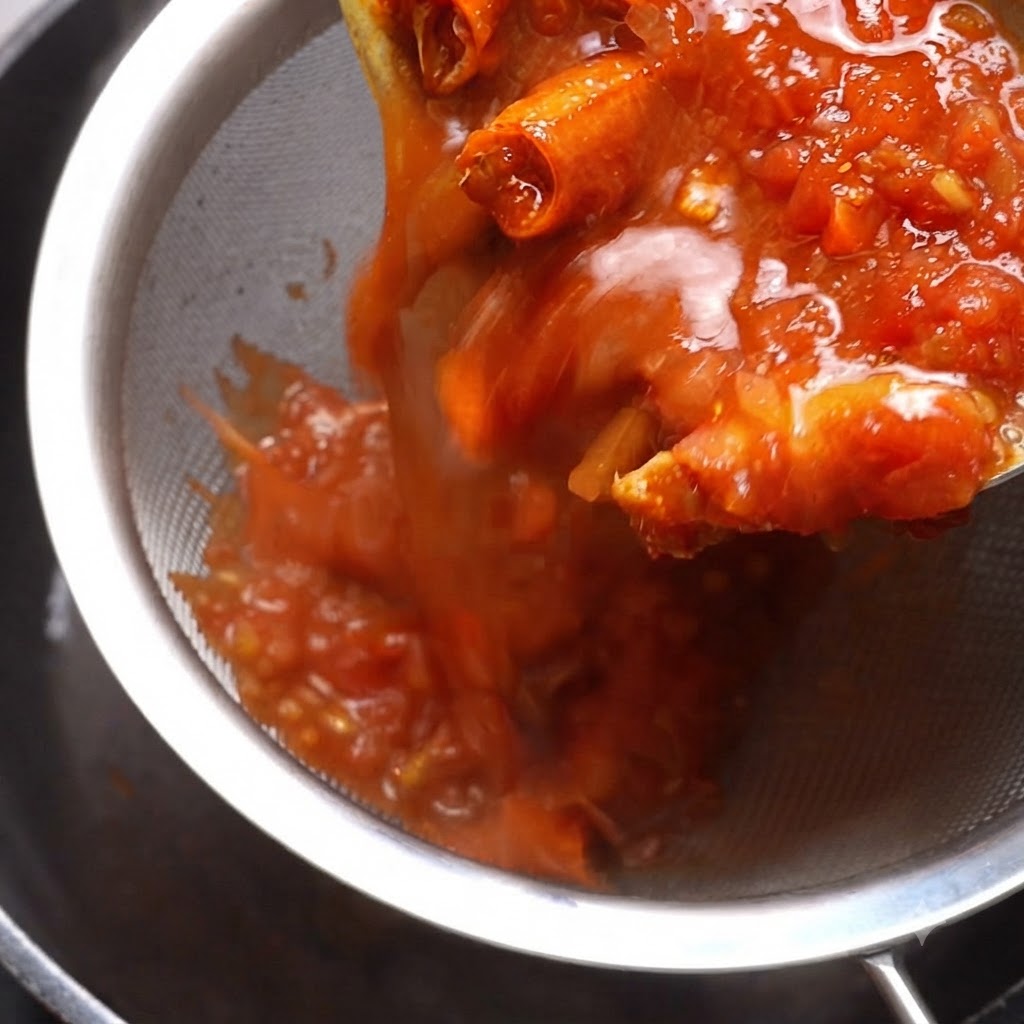 Gekochte Tomatensoße wird durch ein Metallsieb gegossen, während ein Spatel den Inhalt nach unten drückt.