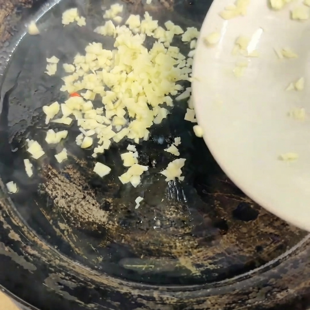 Fein gehackter Knoblauch wird von einem weißen Teller in eine heiße Pfanne gegeben.