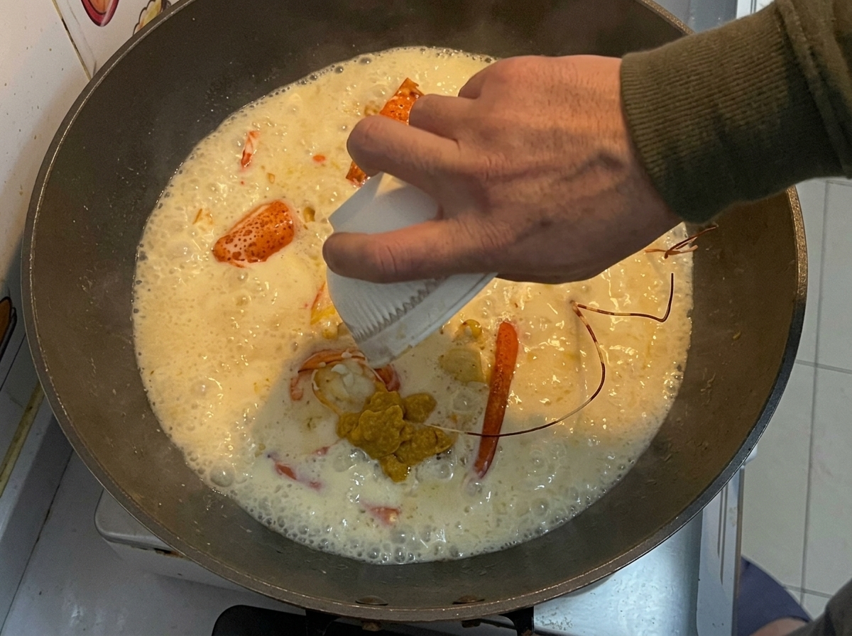Die zurückbehaltene gelbe Langustenpaste wird aus einer kleinen weißen Schüssel in einen Wok mit gebratenen Langustenstücken und einer cremigen Sauce gegossen.