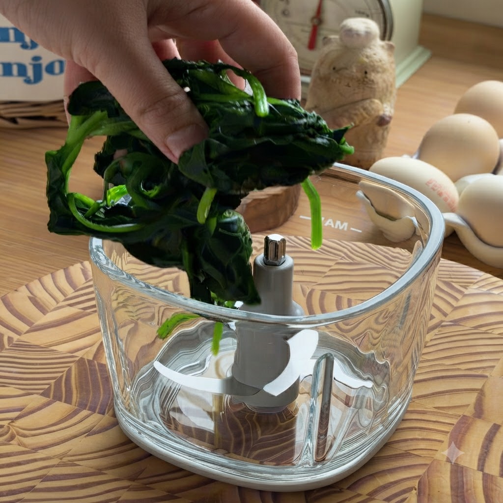 Eine Hand legt einen Klumpen ausgedrückten, gekochten Spinat in eine leere Glas-Küchenmaschinenschüssel.