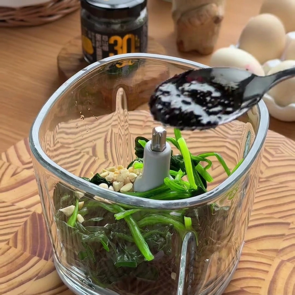 Ein schwarzer Löffel lässt dunkle, reichhaltige schwarze Trüffelsauce in eine mit Spinat und Nüssen gefüllte Küchenmaschinenschüssel tropfen.