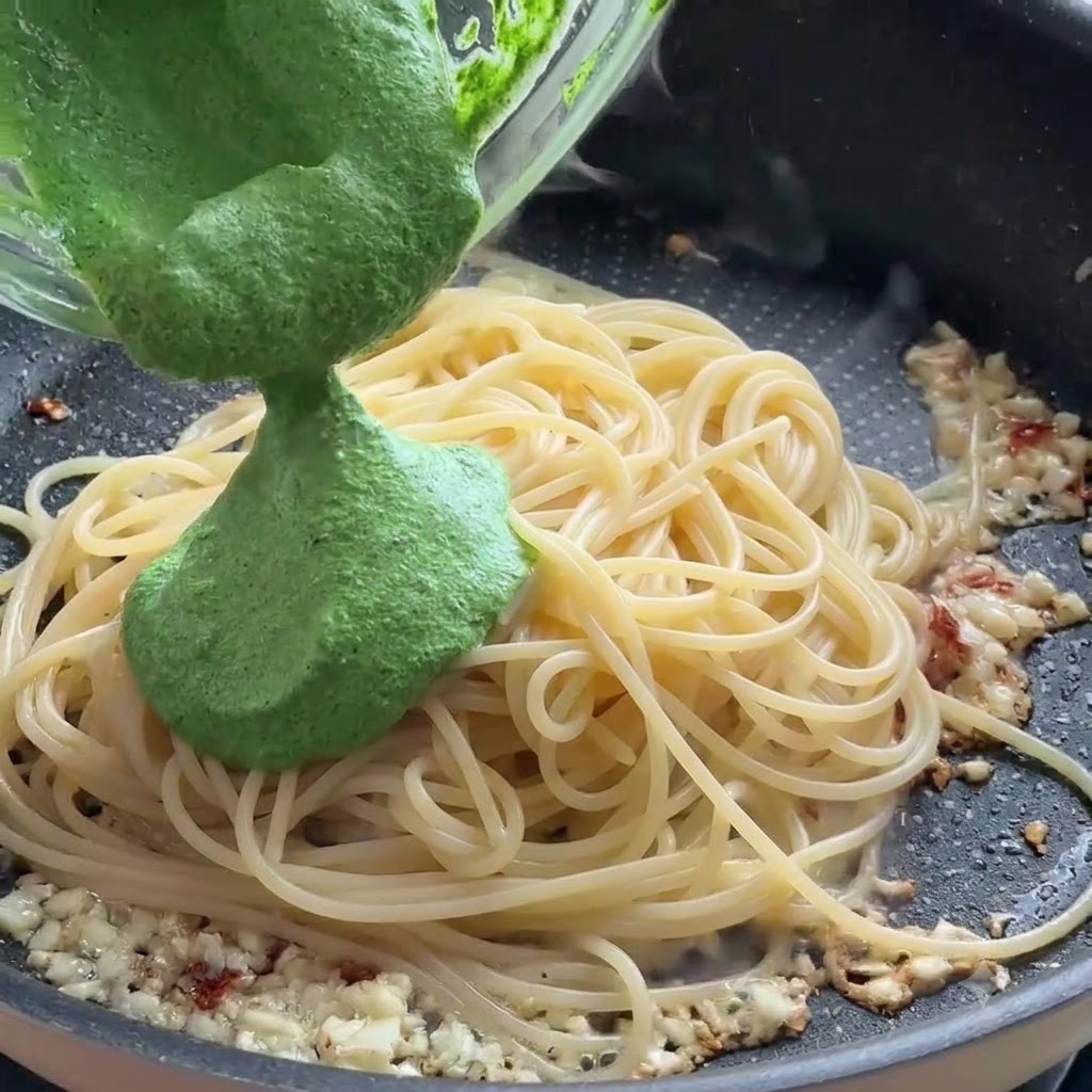 Grüne Pestosauce wird aus einer Glasschüssel auf Spaghetti-Nudeln in einer Bratpfanne gegossen.