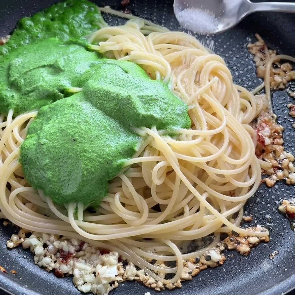 Ein Löffel Salz wird über Spaghetti-Nudeln und grüne Pestosauce in einer Pfanne gestreut.
