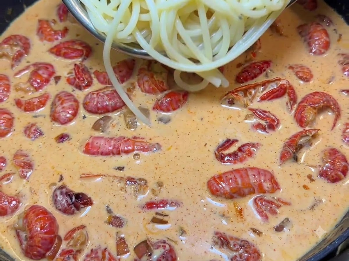 Eine Zange hebt gekochte Spaghetti-Nudeln in eine zischende Pfanne voller cremiger rosa Sauce und roter Krebsschwänze.