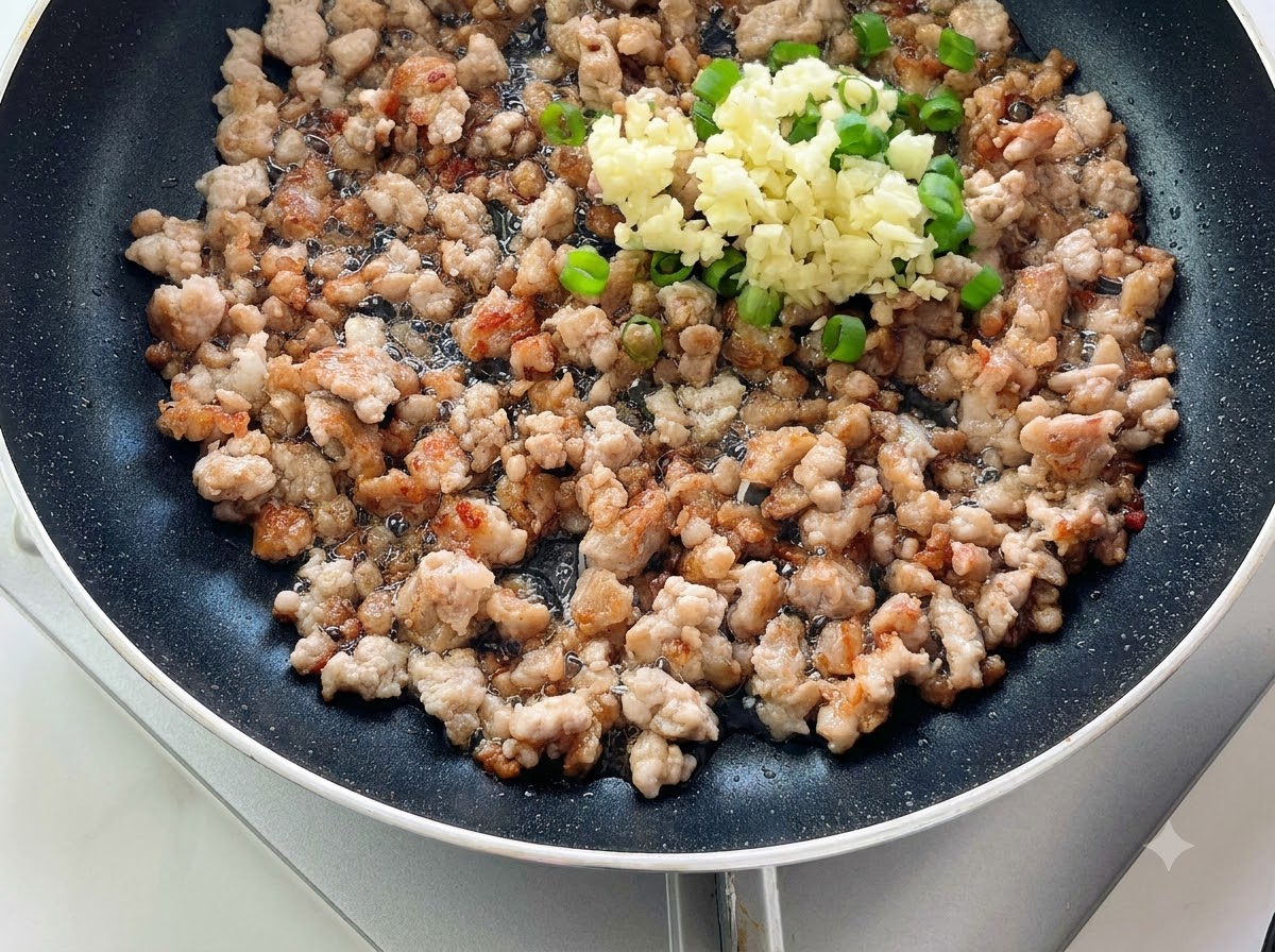 Gehackter Knoblauch und grüne Zwiebeln liegen auf dem gekochten Hackfleisch in einer Pfanne.