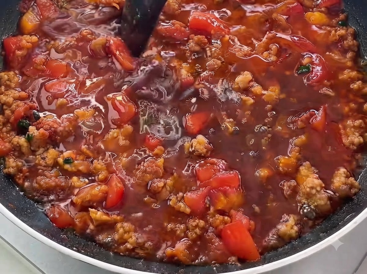 Eine Nahaufnahme dunkler Flüsigkeit, die in eine Pfanne mit angebratenem Hackfleisch und gewürfelten Tomaten gegossen wird.
