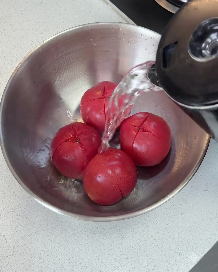 Heißes Wasser wird aus einem Wasserkocher über die eingeschnittenen Tomaten in einer Metallschüssel gegossen.