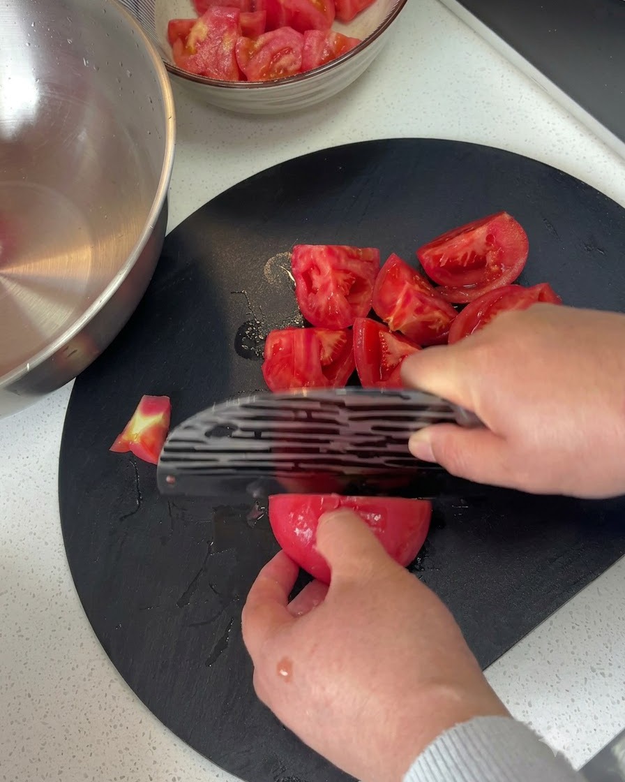 Geschälte Tomaten werden auf einem dunklen Schneidebrett mit einem Messer in Stücke geschnitten.