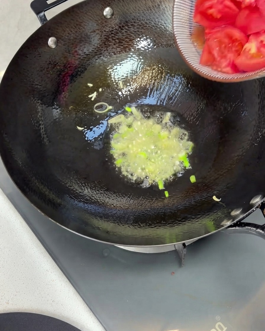 Tomatenstücke werden aus einer Schüssel in einen Wok mit angebratenen Frühlingszwiebeln und Öl geschüttet.