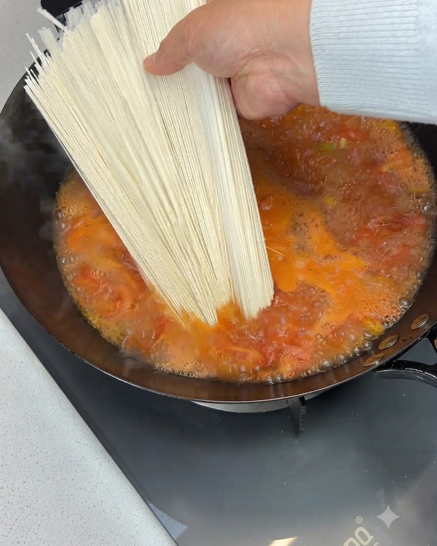Ein Bündel trockener Nudeln wird über eine kochende Tomatensuppe in einem Wok gehalten und hineingegeben.