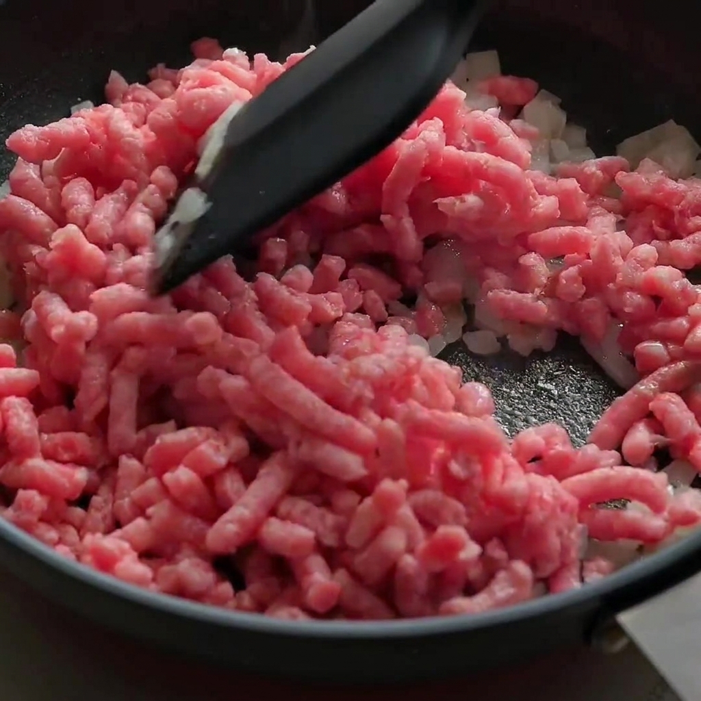 Rohes, rosafarbenes Rinderhackfleisch wird in einer schwarzen Pfanne zerkleinert und mit Zwiebeln angebraten.