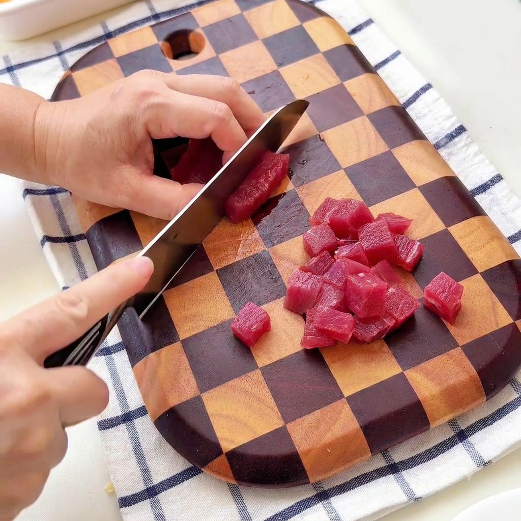 Eine Person schneidet Rindfleisch auf einem karierten Holzschneidebrett in kleine Würfel.