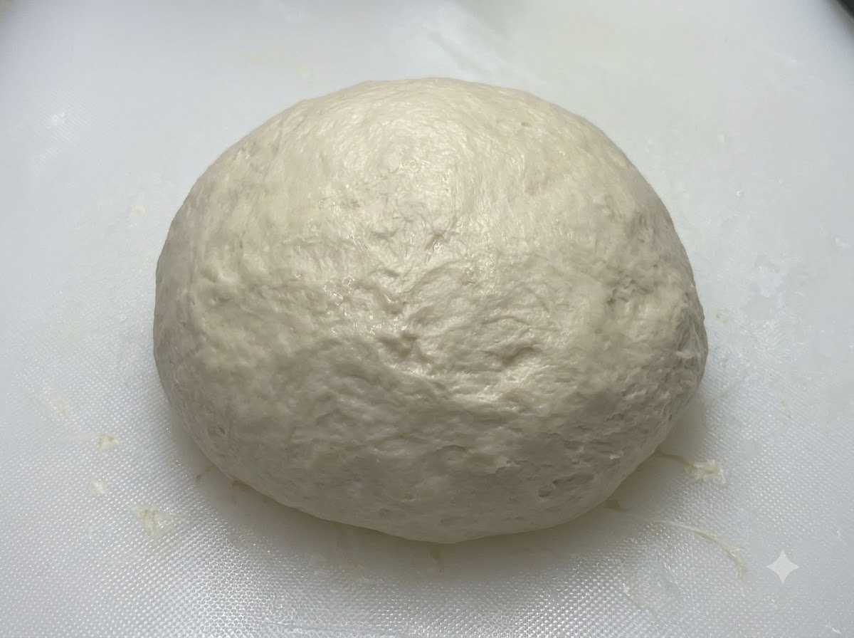 Eine glatte, runde Teigkugel ruht auf einem weißen Brett, bevor sie abgedeckt wird.