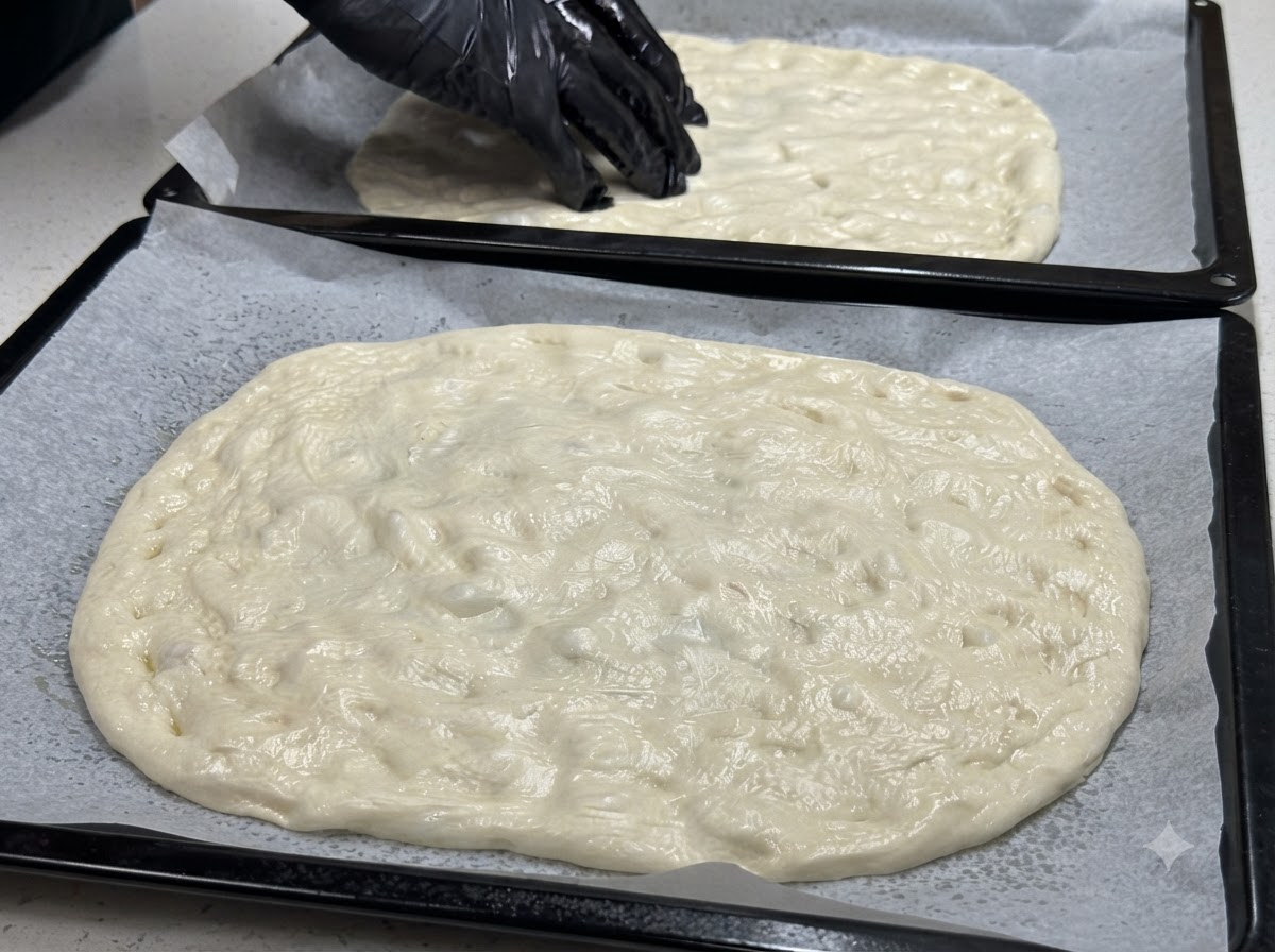 Zwei Backbleche mit flachgedrücktem Teig, auf dem eine behandschuhte Hand halbierte Kirschtomaten verteilt.