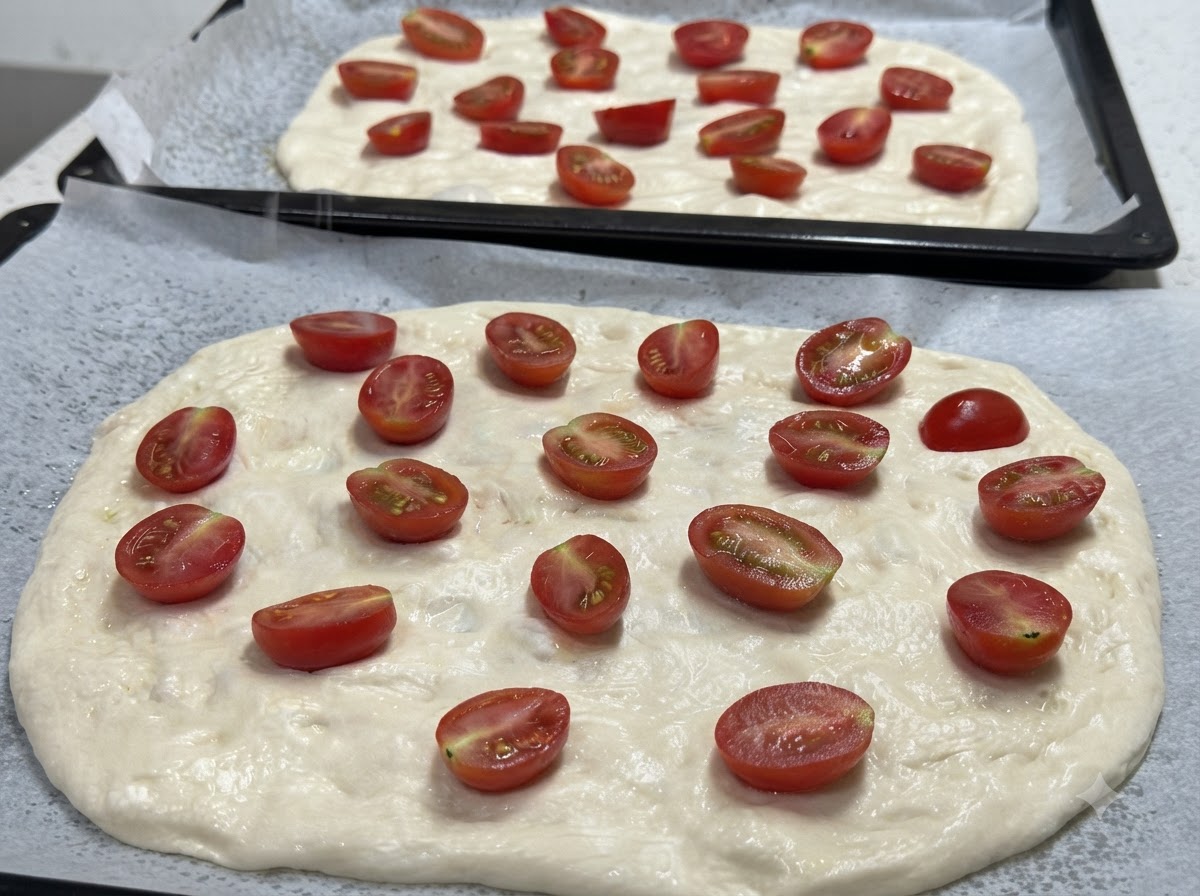 Eine Nahaufnahme von zwei ausgezogenen Pizzaböden, belegt mit halbierten Kirschtomaten, bereit zum Würzen.