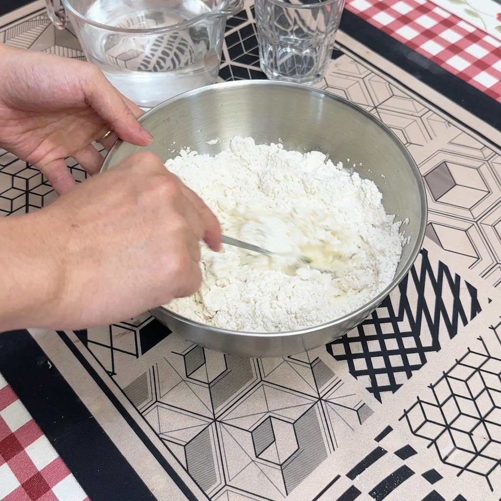 Eine Hand rührt mit einem Löffel Wasser in das trockene Mehl in einer Metallrührschüssel.