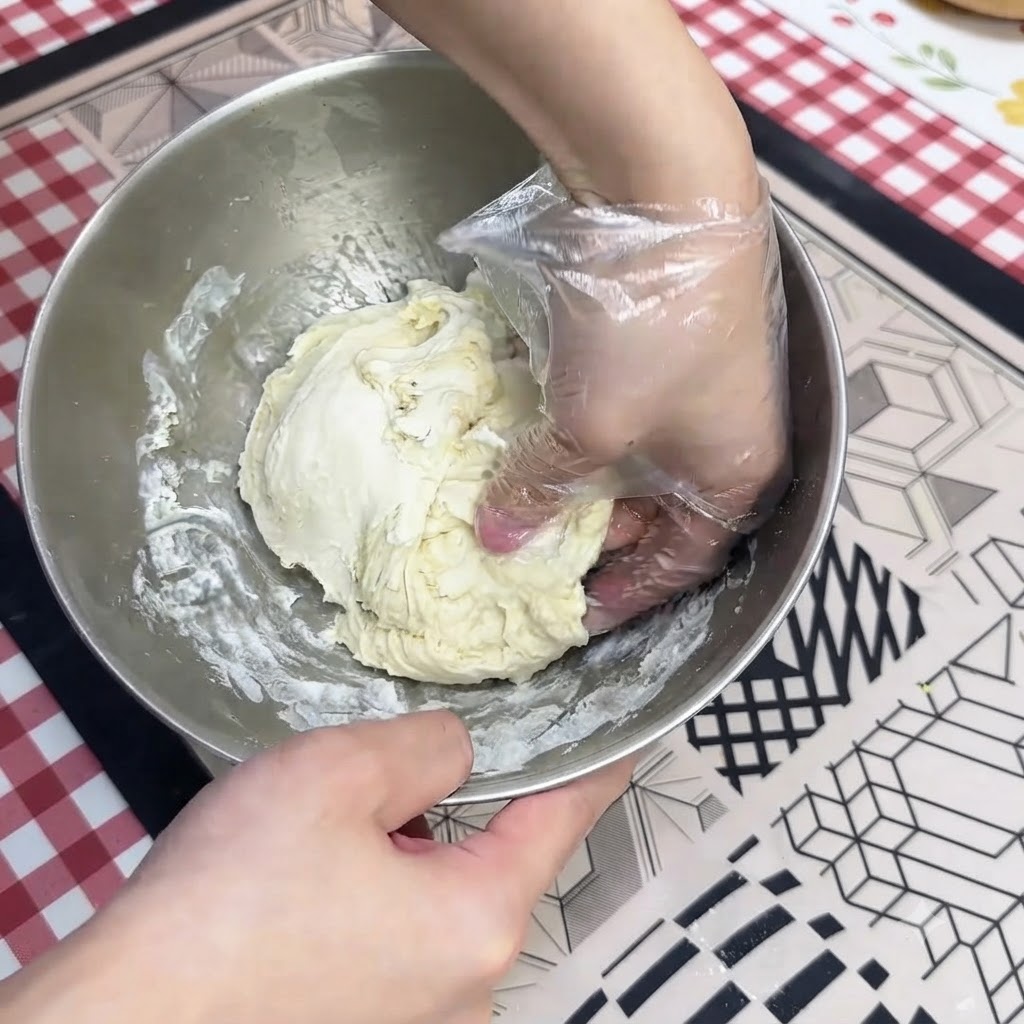 Eine handgeschützte Hand knetet nassen Pizzateig in einer Edelstahlschüssel.