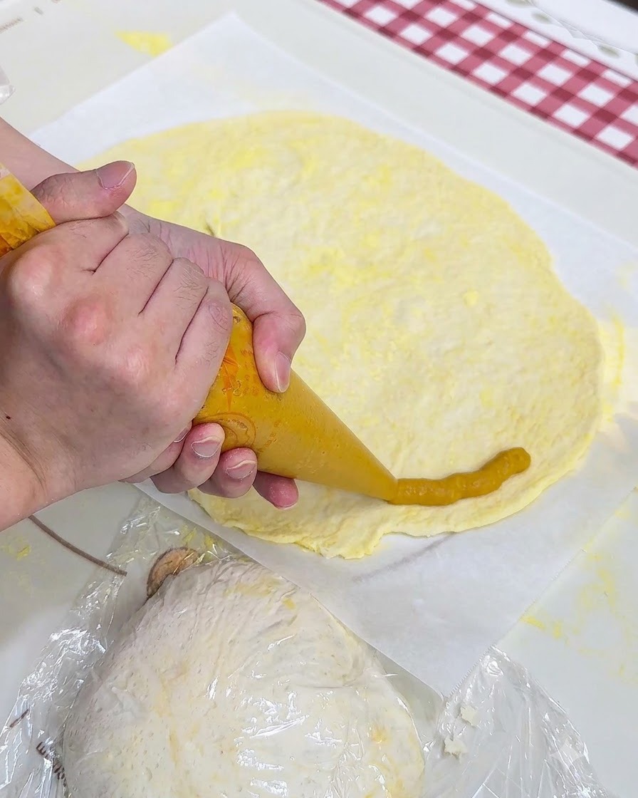 Hände spritzen gelbes Süßkartoffelpüree mit einem Spritzbeutel um den Rand einer flachen Pizzateigplatte auf Backpapier.