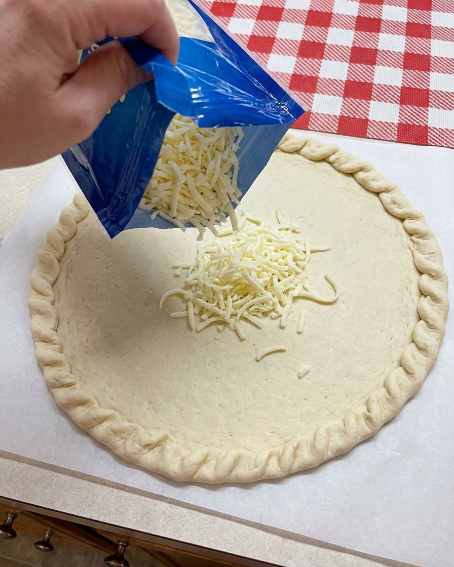 Geraspelter Mozzarella-Käse wird aus einem blauen Beutel auf die Mitte eines vorbereiteten Pizzateigbodens gestreut.