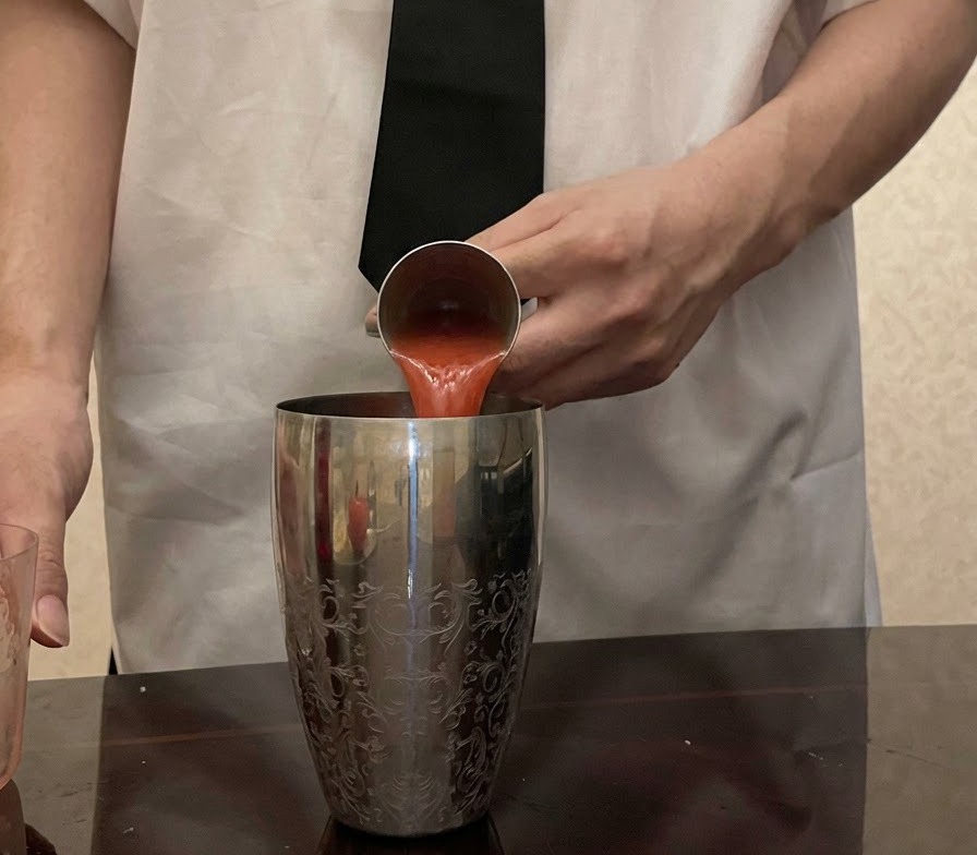 Frischer roter Tomatensaft wird aus einem Jigger aus Metall in einen gemusterten silbernen Cocktailshaker gegossen.