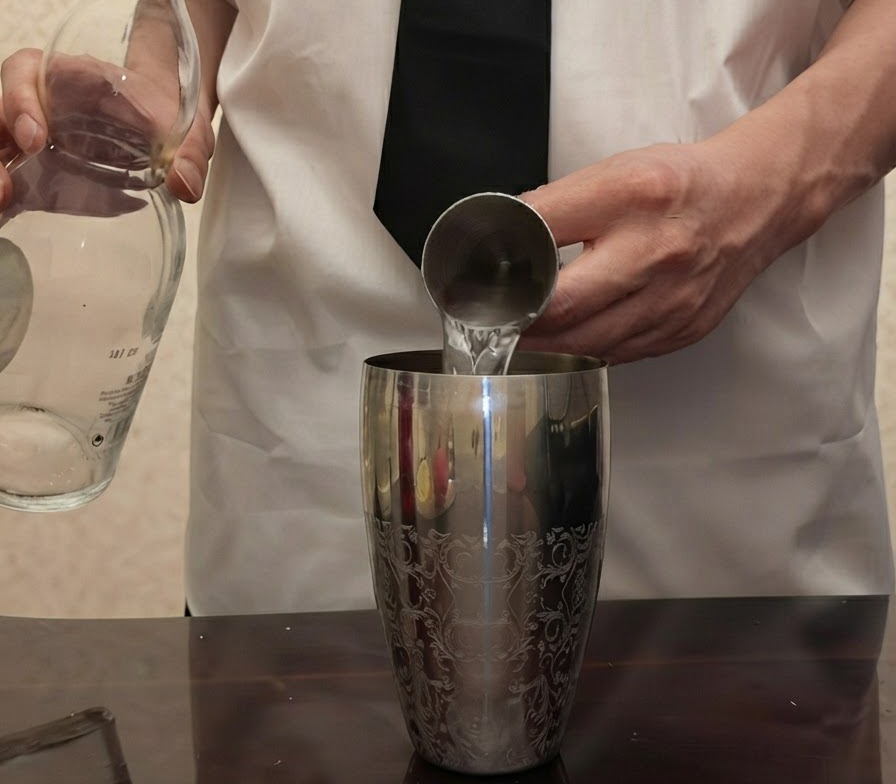 Klarer Tequila wird aus einer Glasflasche in einen über einem silbernen Cocktailshaker gehaltenen Metalljigger gegossen.