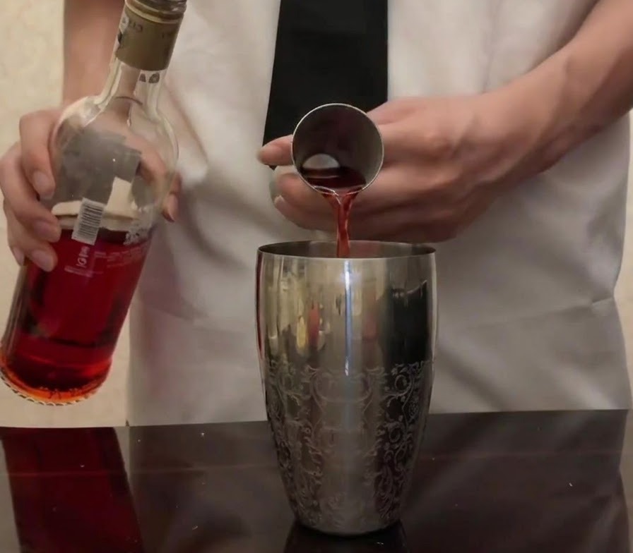 Hellroter Campari wird aus seiner Flasche in einen Metalljigger über einem silbernen Cocktailshaker gegossen.