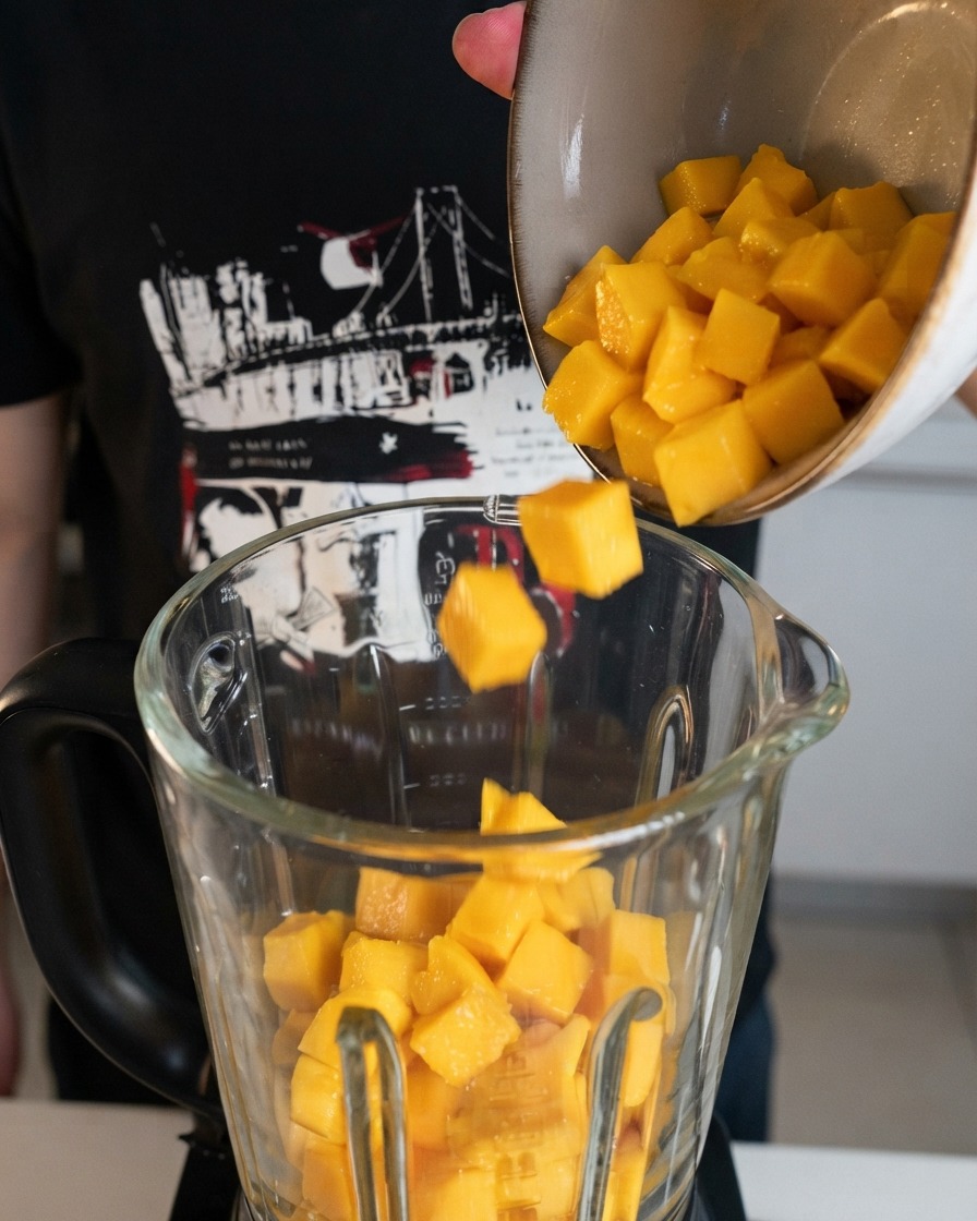 Frisch geschnittene gelbe Mangowürfel werden aus einer Schüssel in einen klaren Glasmixerbehälter geschüttet.