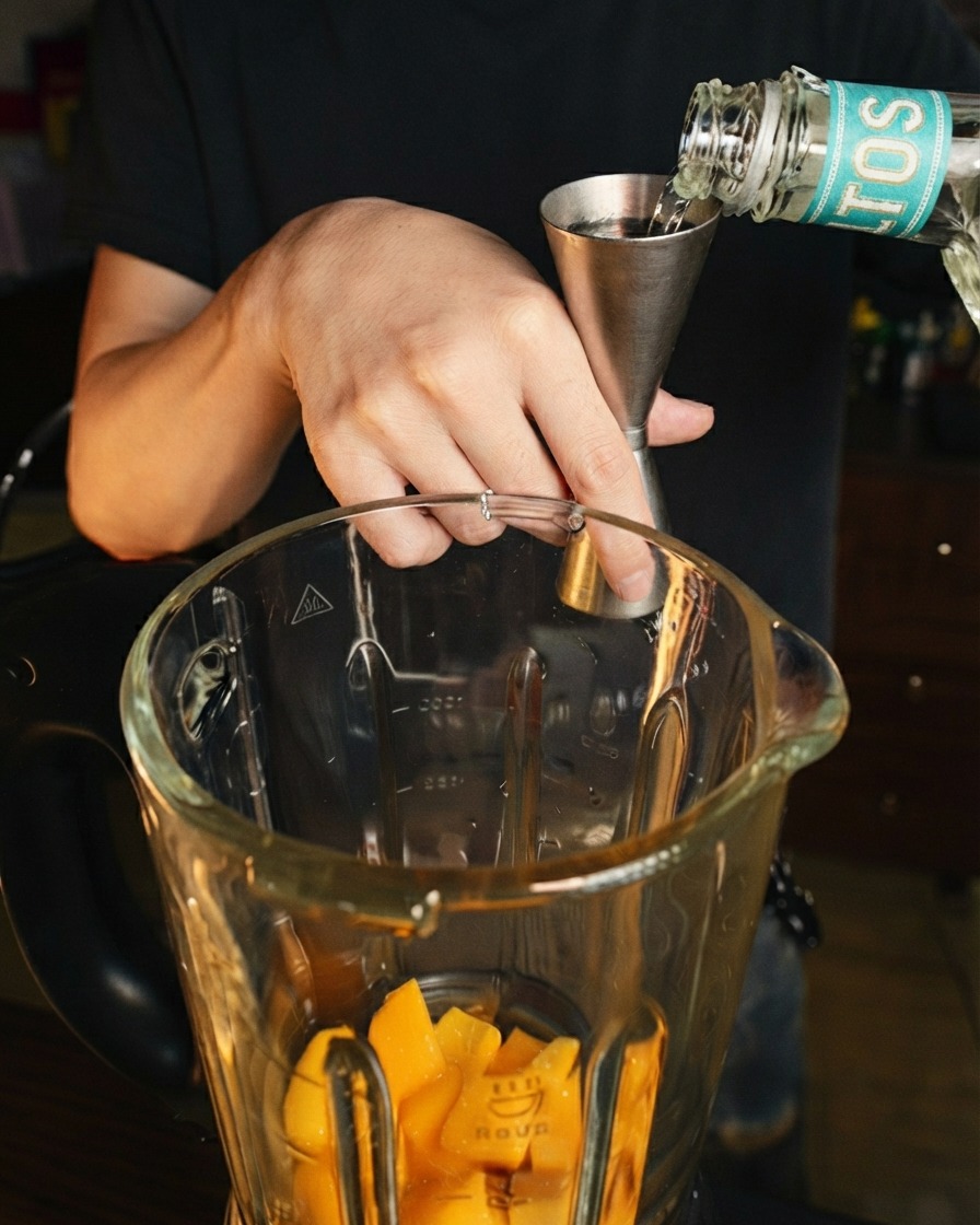 Klarer Likör wird aus einem Edelstahl-Jigger in einen Glasmixbecher über frische Mangostücke gegossen.