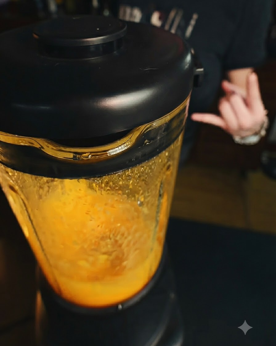 Ein Mixer arbeitet mit einer leuchtend orangen Mangomischung im Inneren, während im Hintergrund eine Hand eine Geste macht.