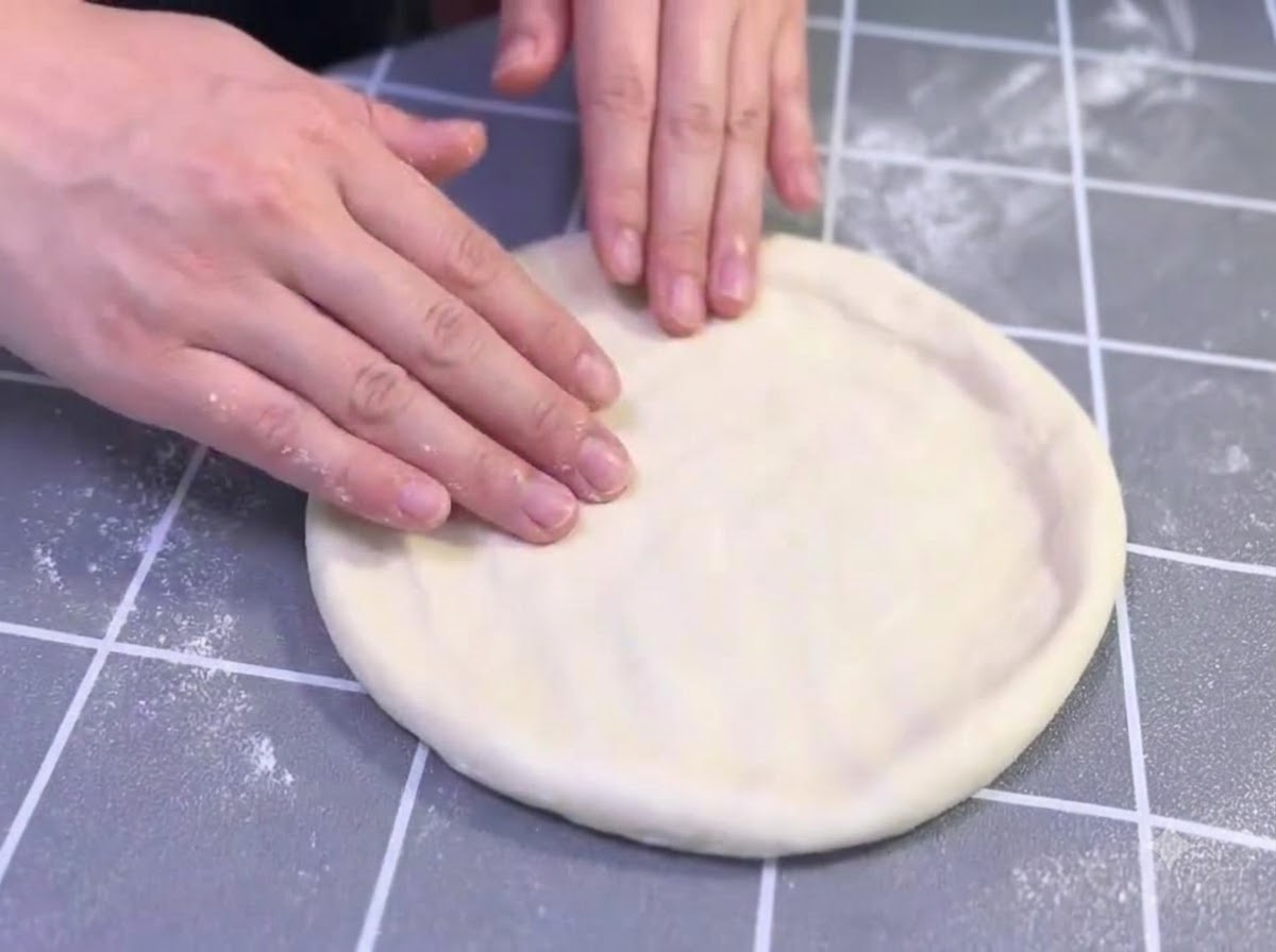Hände dehnen und formen sanft einen geölten Pizzateigling auf einer grauen Rasteroberfläche.
