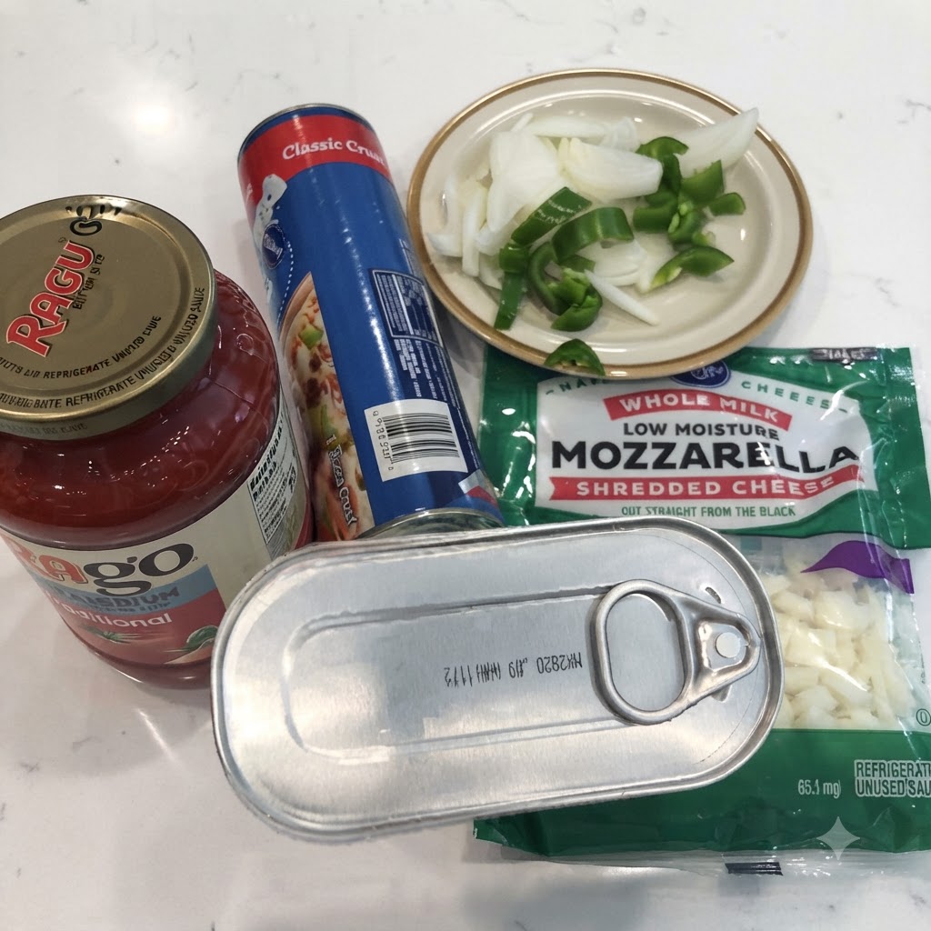 Eine Dose Sardellen, ein Glas Marinarasauce, Pizzateig aus der Dose, geriebener Mozzarella sowie geschnittene Zwiebeln und Paprika auf einer weißen Arbeitsplatte.