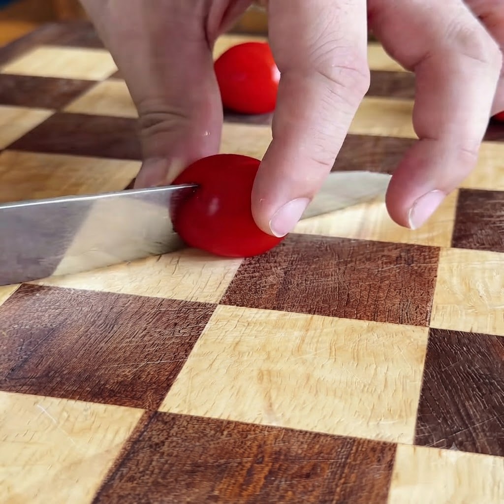 Hände schneiden eine rote Kirschtomate mit einem Messer auf einem karierten Schneidebrett in Hälften.
