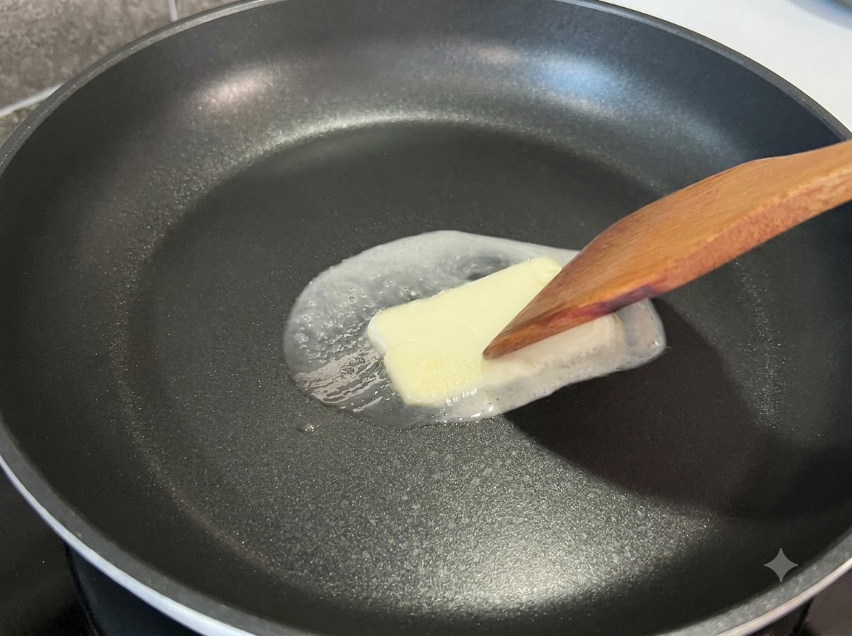 Ein Holzspatel drückt leicht auf ein quadratisches Stück schmelzender Butter in einer schwarzen Bratpfanne.