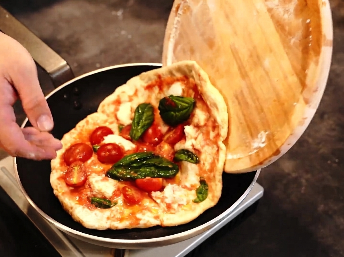 Eine Pizza wird von einem Holzschieber in eine Bratpfanne befördert, während ein Flambierbrenner bereitliegt.