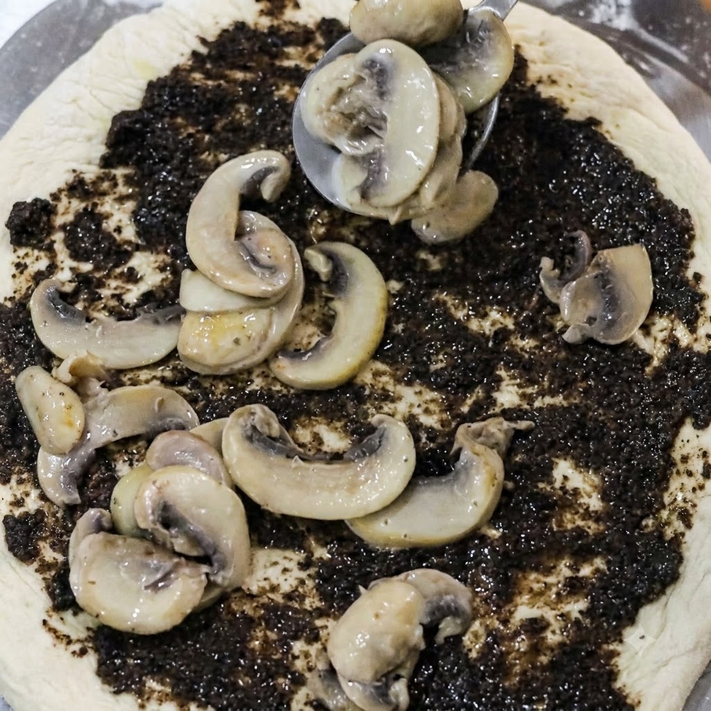 Ein Löffel platziert gekochte Pilzscheiben auf einem mit schwarzer Trüffelpaste bestrichenen Pizzaboden.