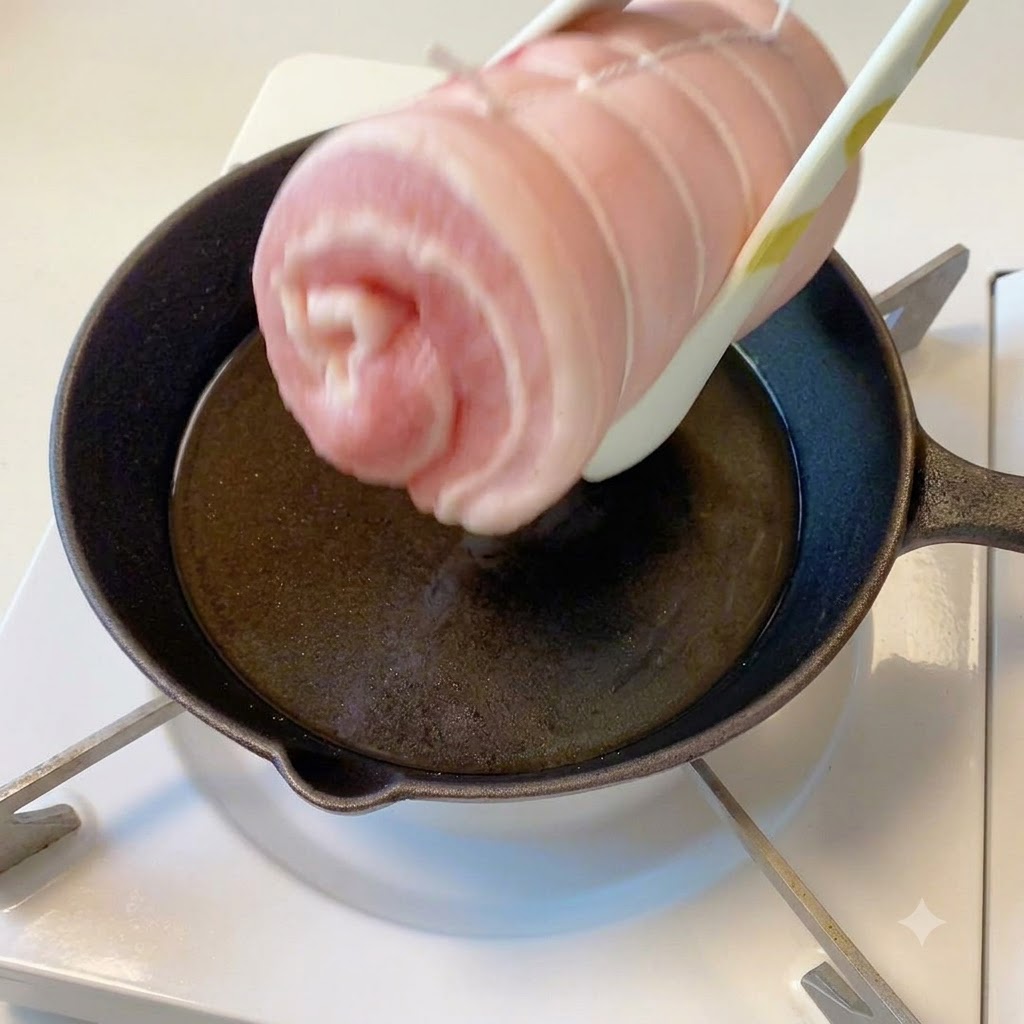 Die gebundene Schweinebauchrolle wird mit einer Zange über einer heißen Pfanne mit Öl gehalten.