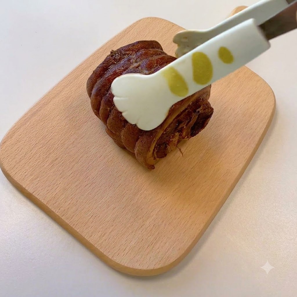 Eine Zange hält ein gekochtes, gerolltes Chashu-Schweinefleischstück auf einem Holzschneidebrett.
