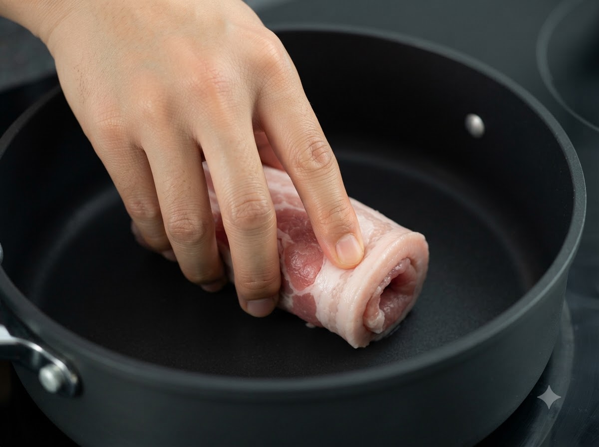 Die verschnürte Schweinebauchrolle wird in einer dunklen Pfanne über Hitze angebraten.