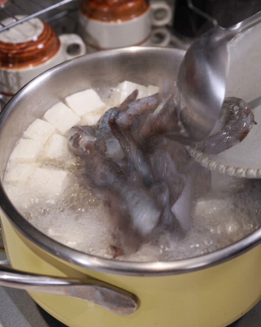 Eine Schöpfkelle gibt rohen Tintenfisch und Garnelenstücke in einen kochenden Suppentopf mit Tofu und Schweinefleisch.