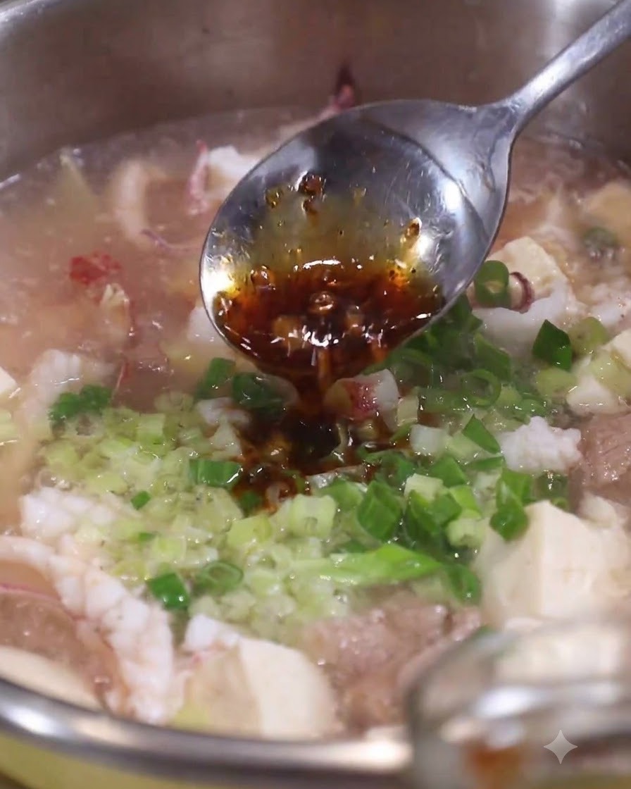 Ein Löffel dunkles Frühlingszwiebelöl wird über frisch gehackte Frühlingszwiebeln und Sellerie in einem Topf mit heißer Meeresfrüchtesuppe geträufelt.
