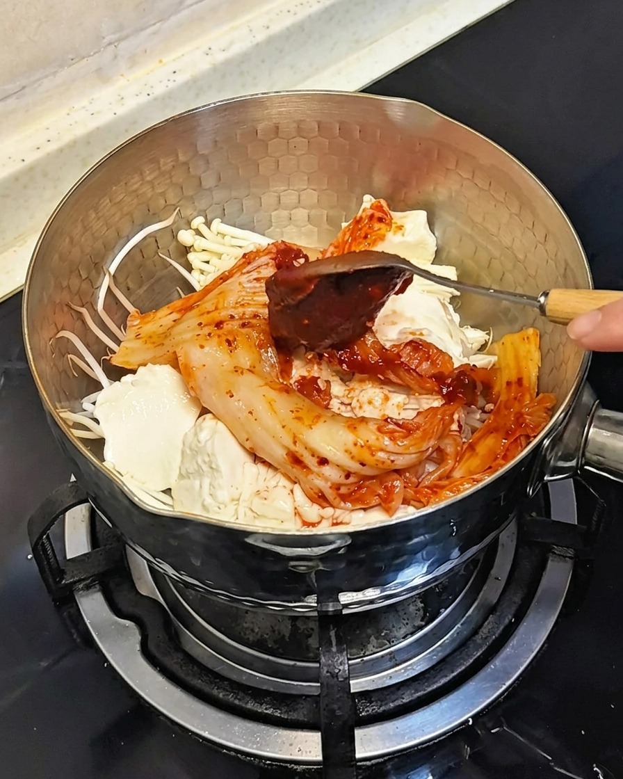 Ein Löffel gibt einen Klecks dicker roter koreanischer Chilispaste auf die Zutaten im Topf.