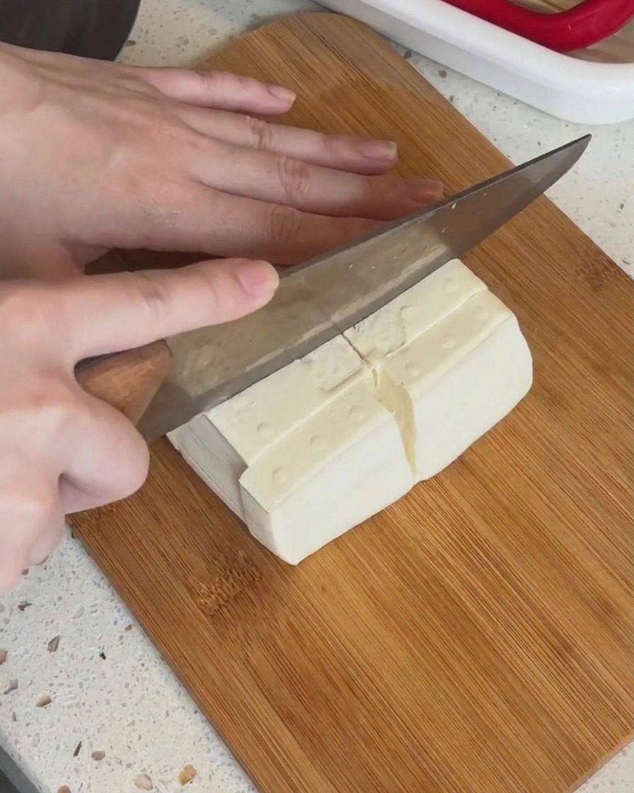 Ein Block weißer Tofu wird auf einem Holzbrett in kleinere rechteckige Stücke geschnitten.