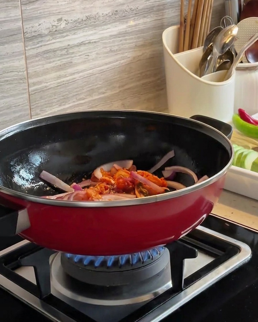 Geschnittene rote Zwiebeln und gehackter Kimchi werden zusammen in einem roten Wok über einer blauen Flamme angebraten.
