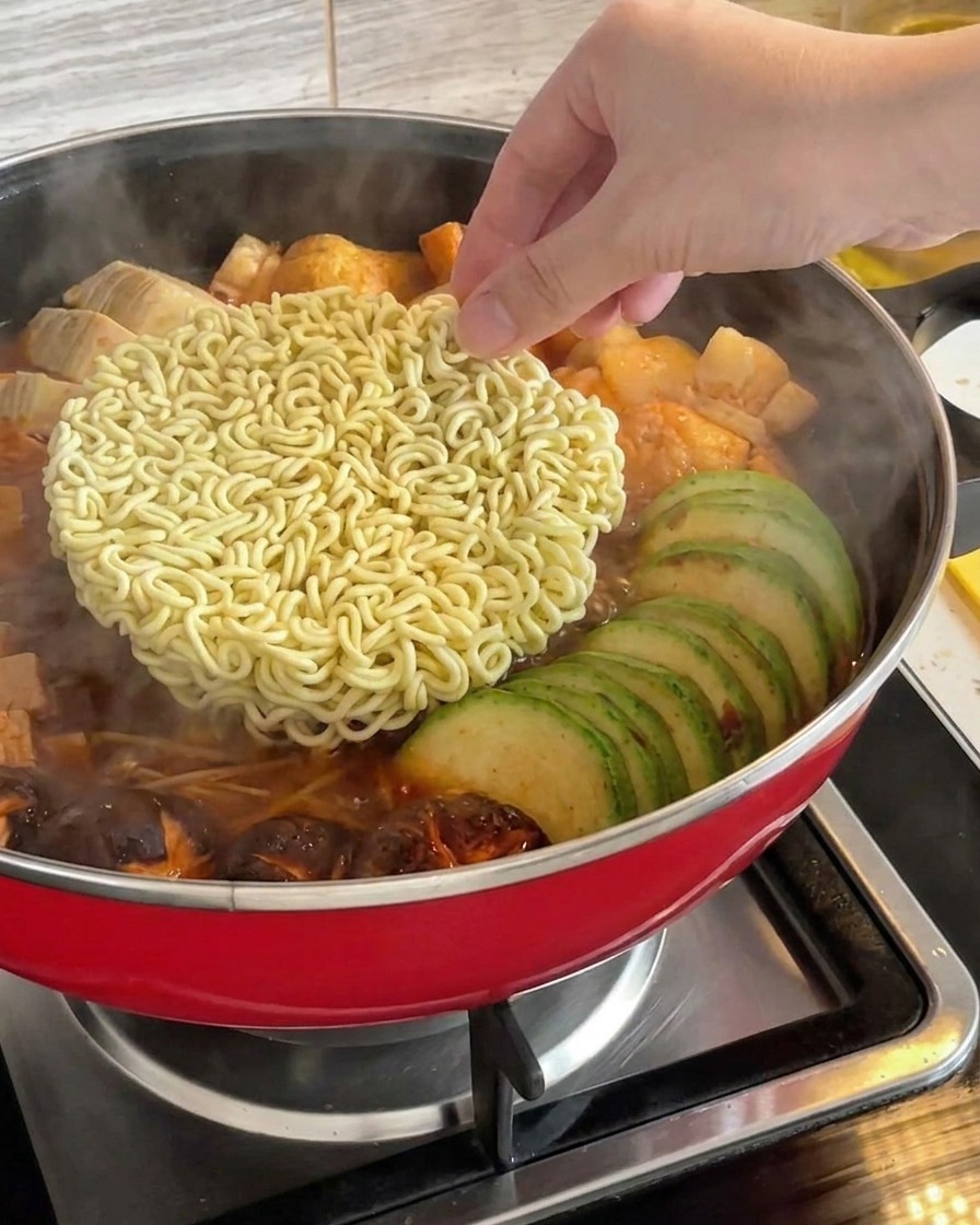 Eine Hand legt einen Block trockene Instant-Ramen-Nudeln in die Mitte eines köchelnden koreanischen Armeeeintopfs.
